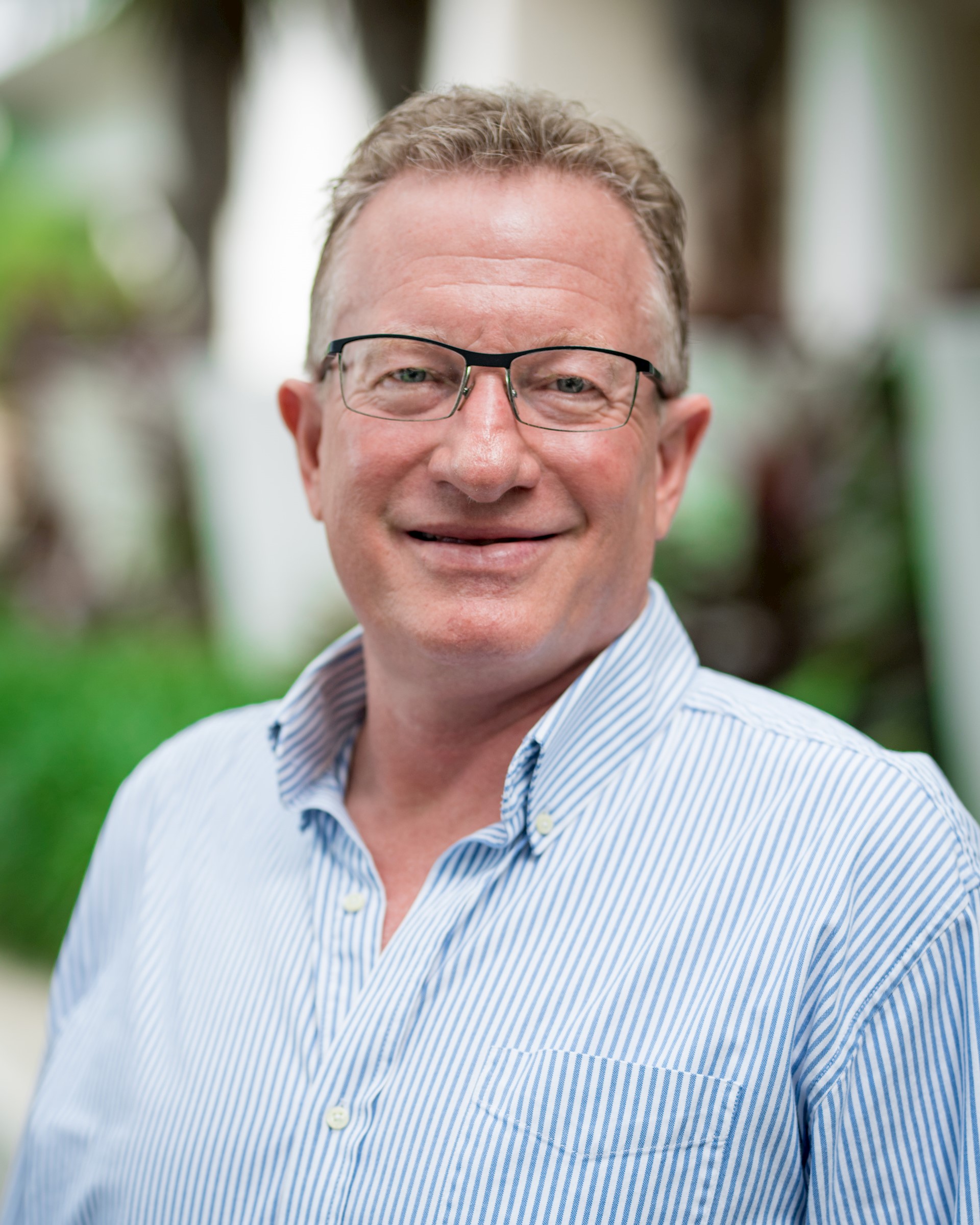 Caucasian man with horn-rimmed glasses wearing a blue shirt smiles at the camera