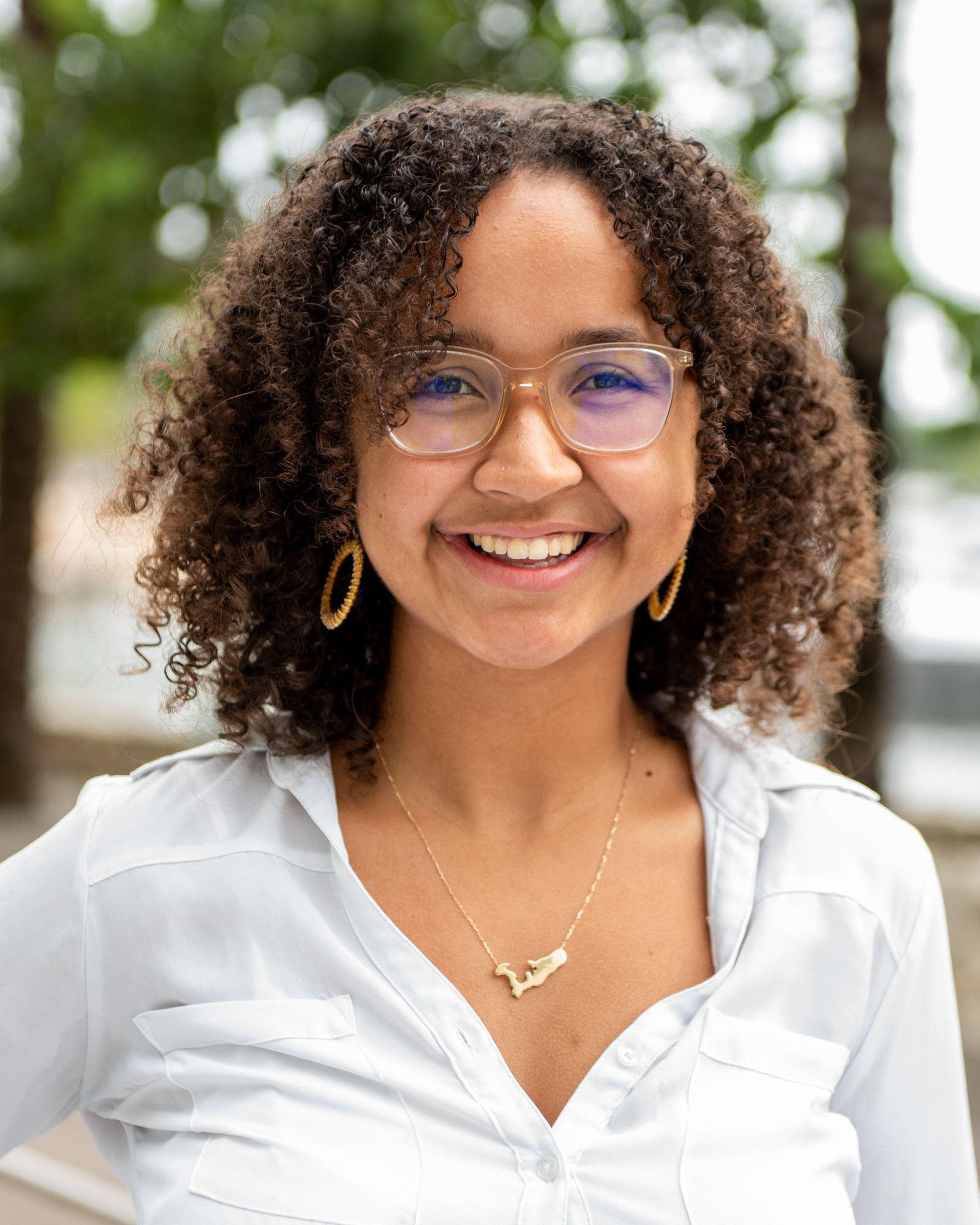 Woman in mid twenties with brown skin and curly brown hair wearing clear glasses smiles at the camera