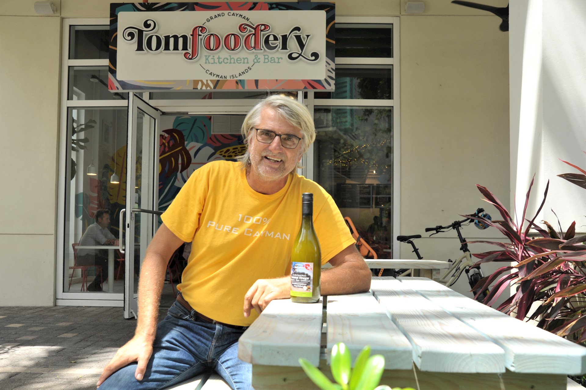 Elder Caucasian man with white hair sitting outside on a bench wearing a yellow shirt smiles at the camera.
