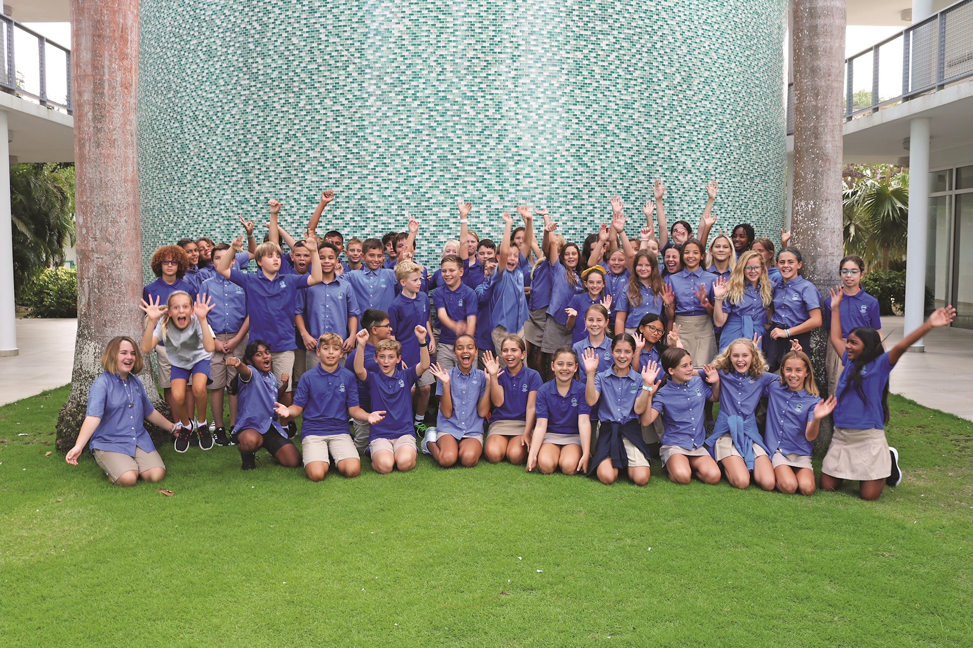 Group of middle schoolers in blue shirts and khaki bottoms smiling while sitting on green grass