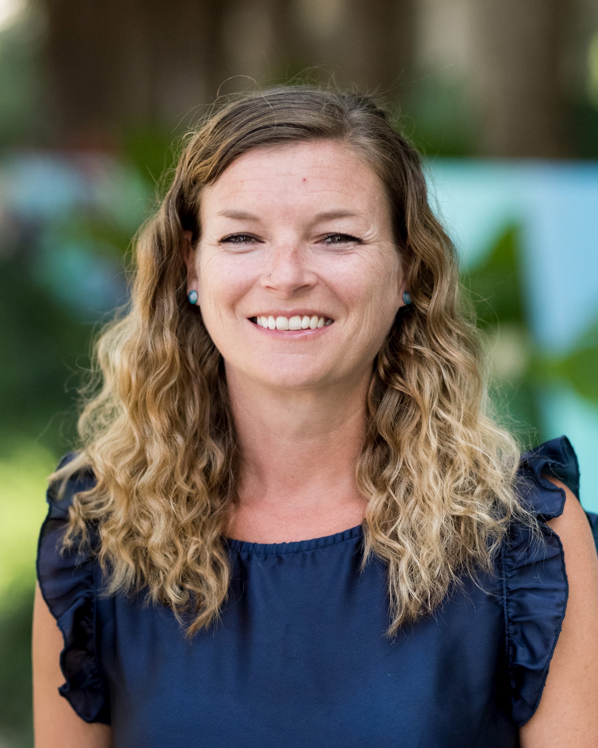 Caucasian woman with dirty blonde wavy hair wearing a navy shirt smiles at the camera