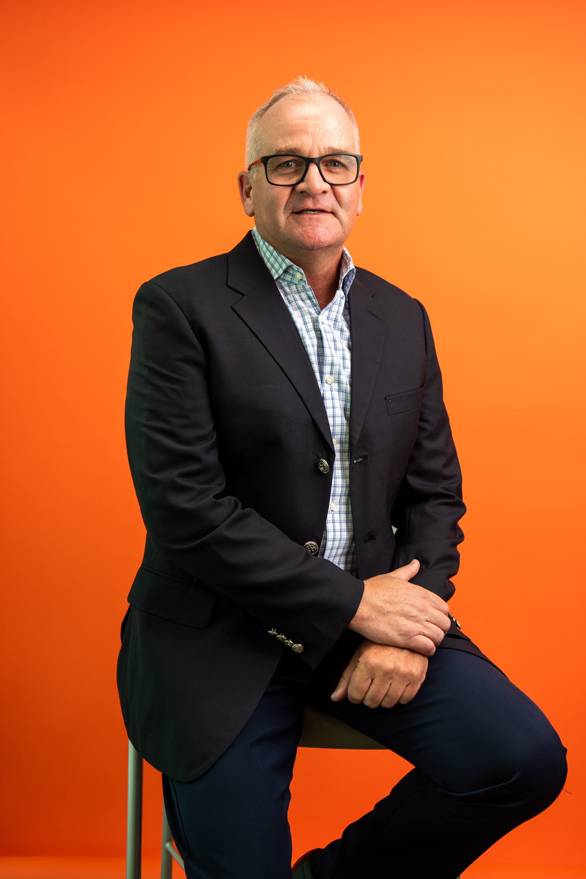 white man with glasses sits on stool smiles at camera